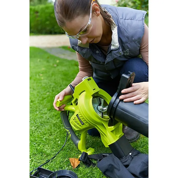 Ryobi Elektro-Laubbläser RBV3000CSV - 3.000 W, Blasgeschwindigkeit: 375 km/h, Blasleistung: 14 m³/min - Frau montiert einen Ryobi Laubbläser im Garten, mit Schutzbrille und Weste. Der Laubsauger ist in Grün und Schwarz gehalten.