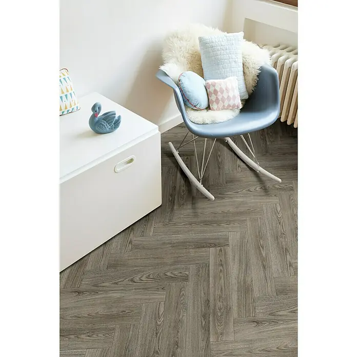 Beauflor PVC-Boden Meterware Hermes - 400 cm, Grau/Braun, Wood 24 - Einladender Raum mit grauem Fischgrätparkett, weißer Kommode, blauem Schaukelstuhl mit Kissen und einem blauen Schwan auf der Kommode.