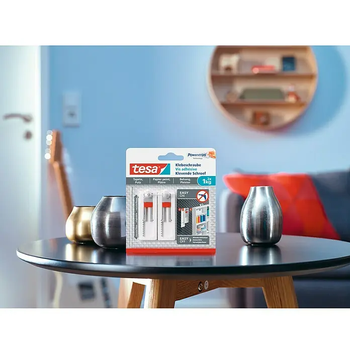 Tesa Tornillo adhesivo ajustable - Rectangular, Carga soportada: 1 kg, 2 ud. - La imagen muestra un paquete de tornillos adhesivos Tesa, colocado sobre una mesa junto con algunos jarrones decorativos.