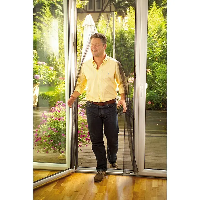 Tesa Insect Stop Mosquitera Comfort - An x Al: 120 x 220 cm, Color red: Antracita - Hombre caminando a través de una puerta con una mosquitera magnética negra, diseñada para evitar la entrada de insectos y dejar entrar aire fresco.