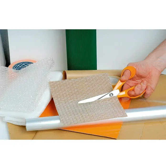 Fiskars Tijera universal Premium - Largo: 21 cm, Acero - La imagen muestra unas tijeras con mangos naranjas cortando un trozo de plástico de burbujas, rodeadas de materiales de embalaje.