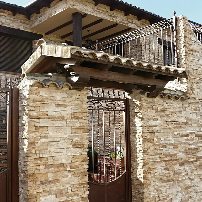 Revestimiento de piedra Petra Arena - Cantidad total: 4 pzs., Arena - Detalle de una entrada con pilares de piedra y una puerta de metal ornamentada, coronada con tejas tradicionales.