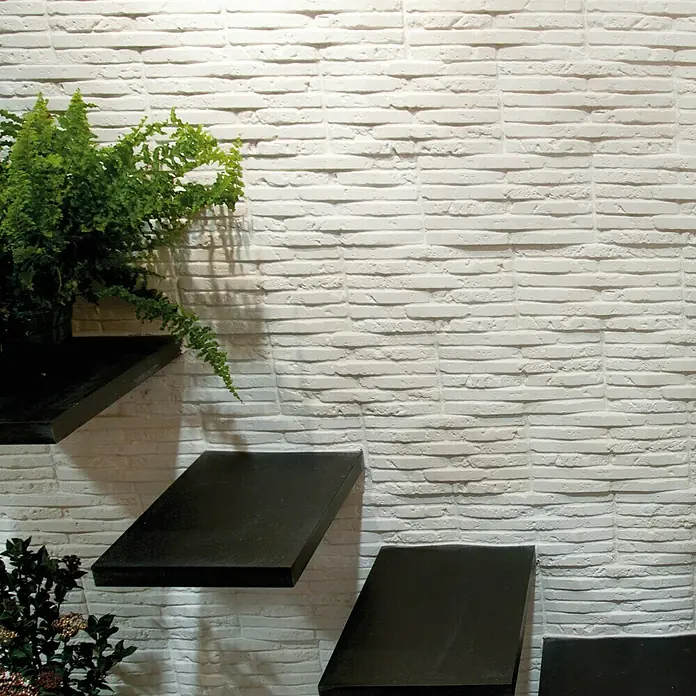 Revestimiento de piedra Listelo Hueso - 28 x 14 x 1 cm - La imagen muestra un fondo de ladrillos blancos con estantes negros y plantas verdes, ofreciendo un elemento de diseño interior elegante.