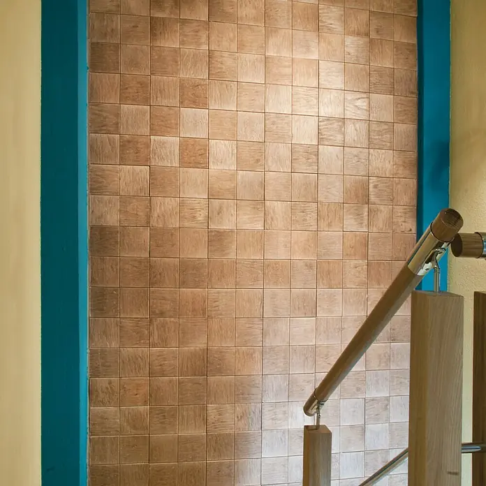 Revestimiento de piedra Teka - Cantidad total: 4 pzs. - Primer plano de una pared con azulejos de madera cuadrados y una barandilla de escalera de madera. Las baldosas son en un tono marrón cálido.