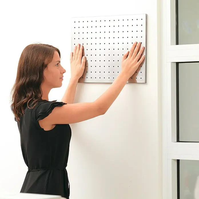 Tesa Powerbond Cinta de doble cara Interior - 19 mm x 5 m - Una mujer fija un tablero perforado cuadrado plateado en una pared blanca, ideal para organizar herramientas.