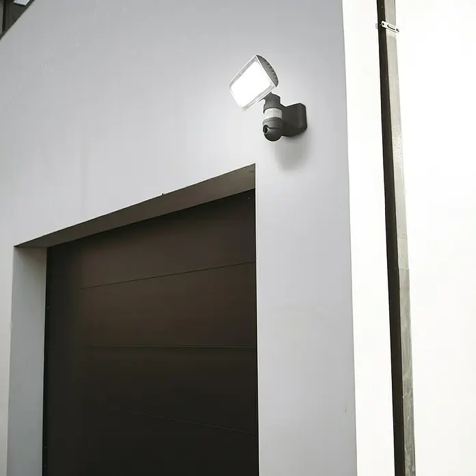 Lutec Proyector LED Peri con sensor y cámara - 25 W, L x An x Al: 22,6 x 20 x 27,1 cm, Gris, Blanco frío - Primer plano de una luz de seguridad LED con cámara montada en una pared blanca sobre una puerta de garaje.