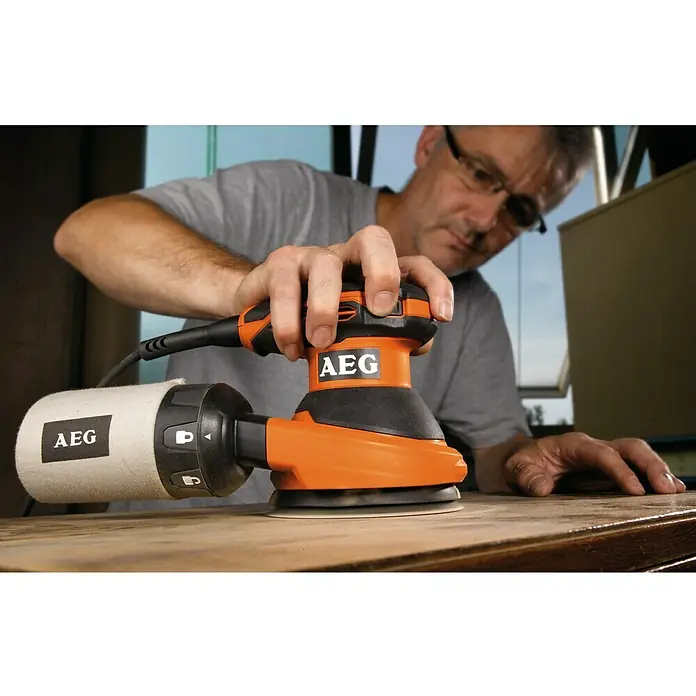 AEG Powertools Elektro-Exzenterschleifer EX 125 ES - 125 mm, 300 W, Leerlaufdrehzahl: 7.000 U/min - 15.000 U/min - Ein Mann benutzt einen AEG Exzenterschleifer, um ein Holzstück zu schleifen. Das orangefarbene Werkzeug liegt flach auf dem Holz.