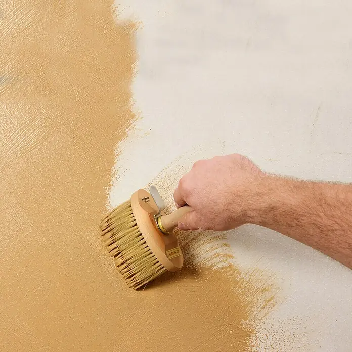 Claytec Lehm-Streichputz Clayfix - Kolumbagrau, 10 kg - Ein Breitpinsel mit Holzgriff wird verwendet, um eine Wand mit beiger Farbe zu streichen, wobei der unbehandelte Teil sichtbar ist.