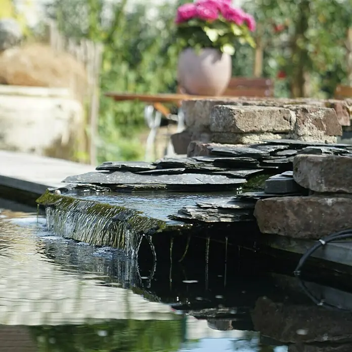 Ubbink Bachlaufpumpe Cascademax 12000 - 100 W, Fördermenge pro Std.: 11.800 l - Nahaufnahme eines kleinen Wasserfalls mit grauen Steinen, der in einen dunklen Teich mündet, mit Pflanzen und einem Tisch im Hintergrund.