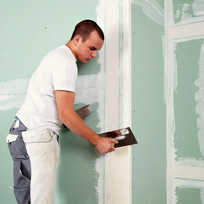 Rigips Kantenschutz für Trockenbau Aqua Bead - 250 cm, Kunststoff - Ein Handwerker glättet mit einer Spachtel die Spachtelmasse an einer Gipskartonwand. Die Wand ist grünlich und uneben.