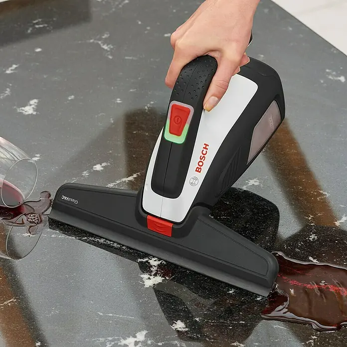 Bosch Akku-Fenstersauger GlassVac Set - 3,6 V, 2 Ah - Das Bild zeigt einen Bosch GlassVac Fensterreiniger, der verschütteten Wein auf einer dunklen Arbeitsfläche aufsaugt. Das Gerät ist weiß und schwarz.