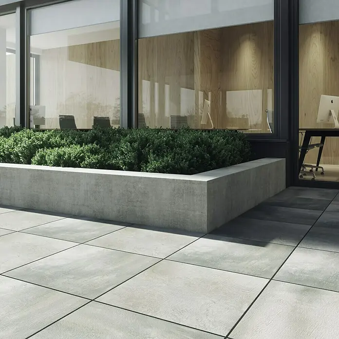Cersanit Terrassenfliese Beton Grey Light - 59,3 x 59,3 x 2 cm, Grau, Matt - Außenaufnahme mit hellgrauen Terrassenfliesen und einem erhöhten Pflanzgefäß mit grünen Büschen vor einem modernen Gebäude mit Fensterfassade.