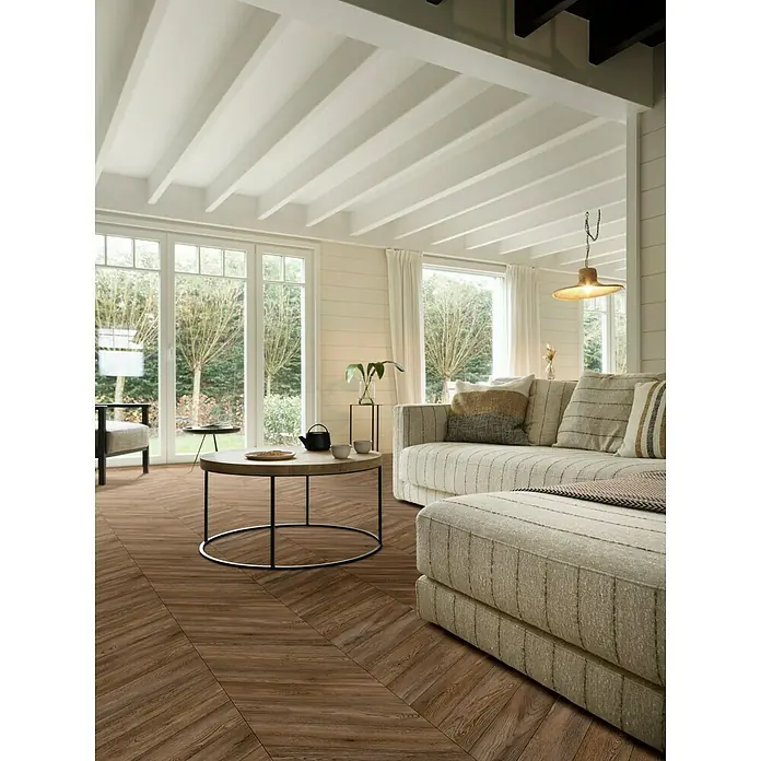 Beauflor PVC-Boden Meterware Calypso - 400 cm, Dunkelbraun - Elegantes Wohnzimmer mit Herringbone-Holzboden, der Wärme und Stil vereint. Die Einrichtung umfasst ein bequemes Sofa und einen runden Couchtisch.
