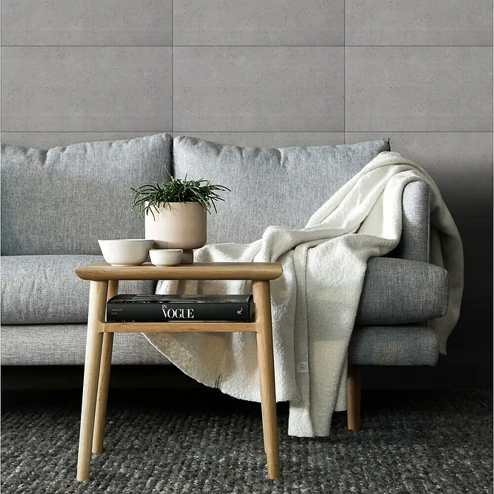 Nmc Decoflair Wandpaneel - Beton Light, 100 x 50 cm, Kunststoff - Ein stilvoller Beistelltisch aus hellem Holz mit Ablage, dekoriert mit einer Vogue-Zeitschrift, Schalen und einer Pflanze, vor einem grauen Sofa.