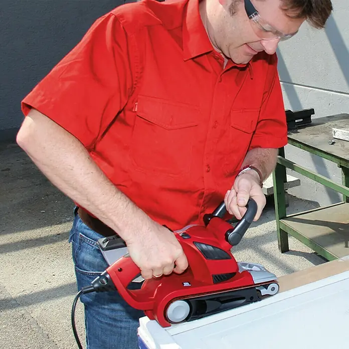 Einhell Expert Lijadora de banda eléctrica TE-BS 8540 E - 850 W - Primer plano de un hombre usando una lijadora de banda roja sobre una pieza de madera. La lijadora es el foco principal de la imagen.