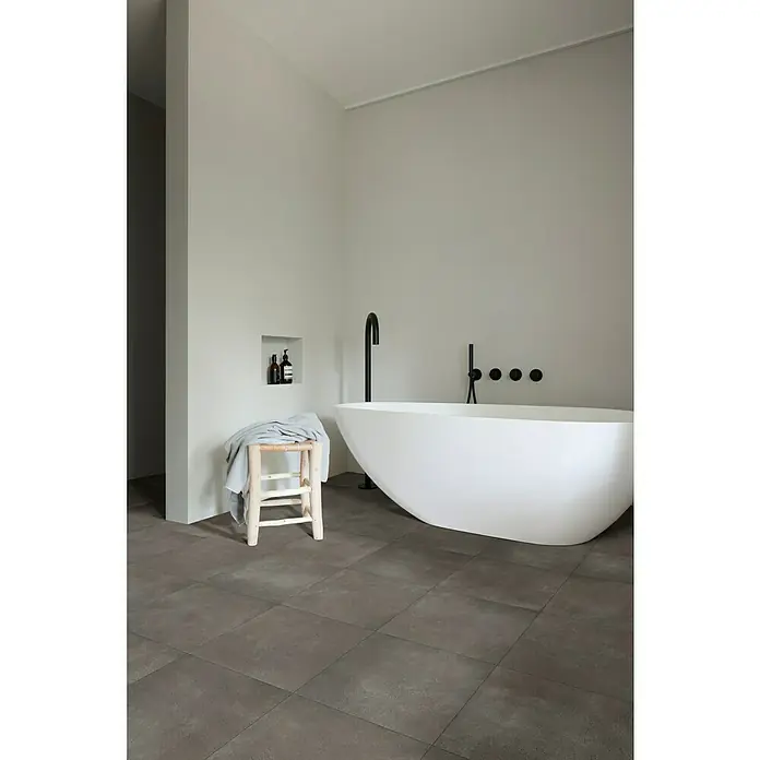 Beauflor PVC-Boden Meterware Jupiter - 400 cm, Dunkelgrau, Tile 23 - Ein stilvolles Badezimmer mit einer freistehenden Badewanne, einem Holzhocker und grauen Bodenfliesen. Das minimalistische Design schafft eine entspannende Atmosphäre.