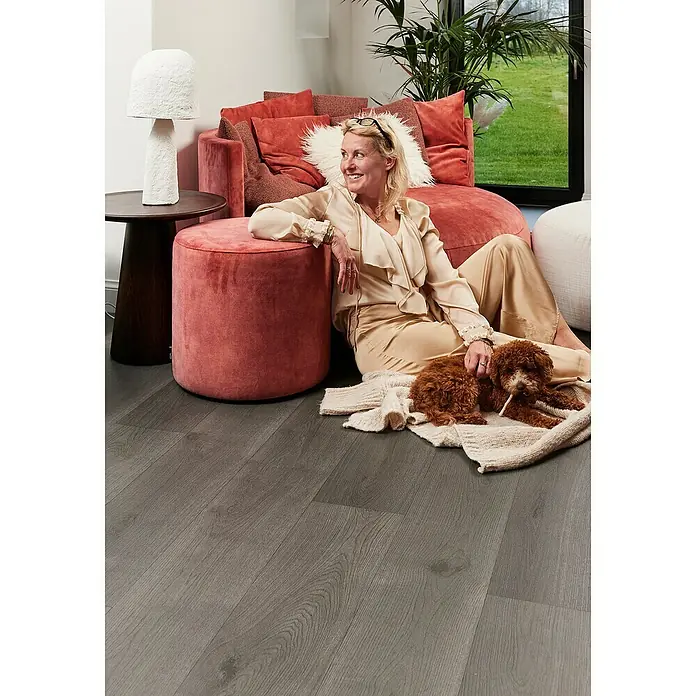 Beauflor PVC-Boden Meterware Jupiter400 cm, Wood 21, Dunkelgrau - Dieses Bild zeigt einen grauen Holzboden mit einer Frau, einem Hocker und zwei Hunden. Der Boden ist der Fokus des Bildes.