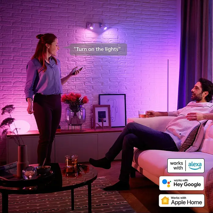 Philips Hue Smart-LED-Lampe White & Color Ambiance - GU10, Dimmbar, RGBW, 400 lm, 4,2 W, 1 Stk. - Intelligente Beleuchtung im Wohnzimmer: Eine Frau steuert die Lampen per Sprachbefehl, während ein Mann entspannt auf der Couch sitzt.