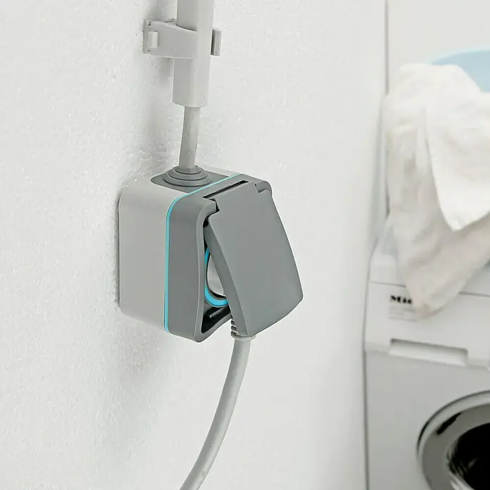 Voltomat Blueline Feuchtraum-Steckdose - 1-fach, Horizontal, IP54, Grau - Eine spritzwassergeschützte Steckdose mit Klappdeckel und einem angeschlossenen Kabel in einer Waschküche, die Sicherheit und Funktionalität vereint.