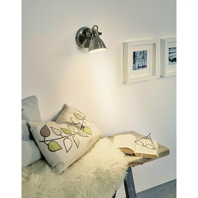 Eglo LED-Wandstrahler Seras - 3,3 W, Creme, Nickel Antik, Warmweiß - Nahaufnahme einer verstellbaren Wandleuchte aus Metall, die über einem gemütlichen Lesebereich mit Kissen und Buch angebracht ist.