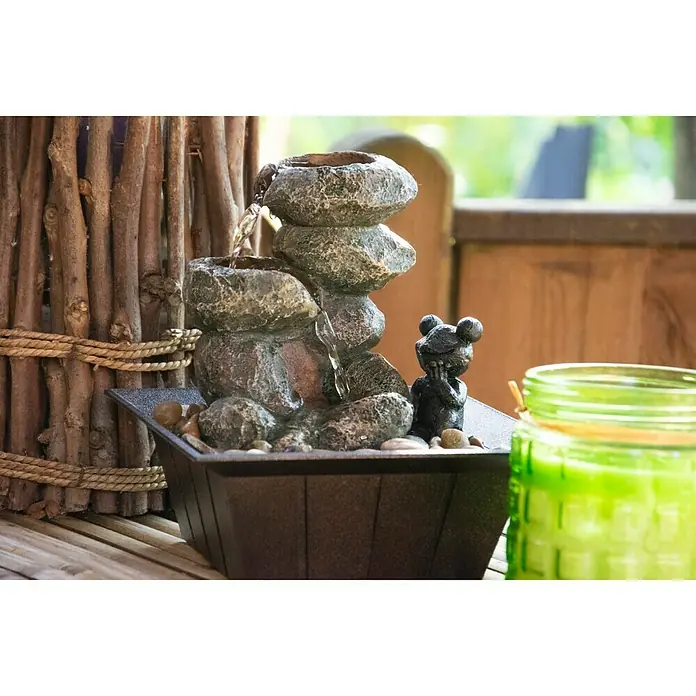 Dobar Gartenbrunnen - 20 x 20 x 21,5 cm, Kunststoff, Grau/Schwarz - Das Bild zeigt einen Zimmerbrunnen mit einer kleinen Froschfigur, umgeben von einem dekorativen Holzzaun und einem grünen Glasbehälter.