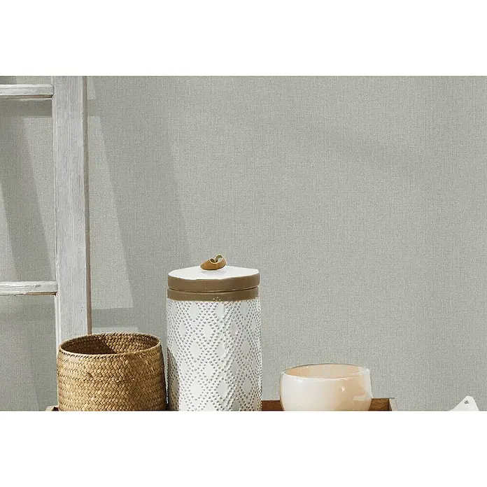 A.S. Création Hygge Vliestapete - Beige, Uni, 10,05 x 0,53 m - Nahaufnahme eines weißen Dekogefäßes mit Deckel, einem gewebten Korb und einer cremefarbenen Schale auf einem Holztisch vor einer neutralen Wand.