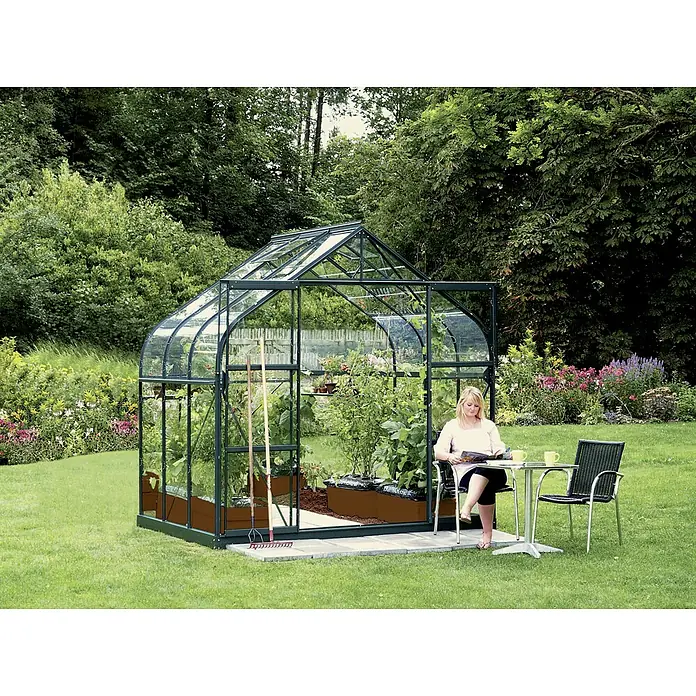 Vitavia Gewächshaus Dione 5000 - Außenmaß inkl. Dachüberstand (B x T): 263,5 x 195 cm, 3 mm, Smaragd - Frau entspannt sich neben dem Gewächshaus. Das Gewächshaus ist mit Pflanzen und Gartengeräten ausgestattet und fügt sich gut in den Garten ein.