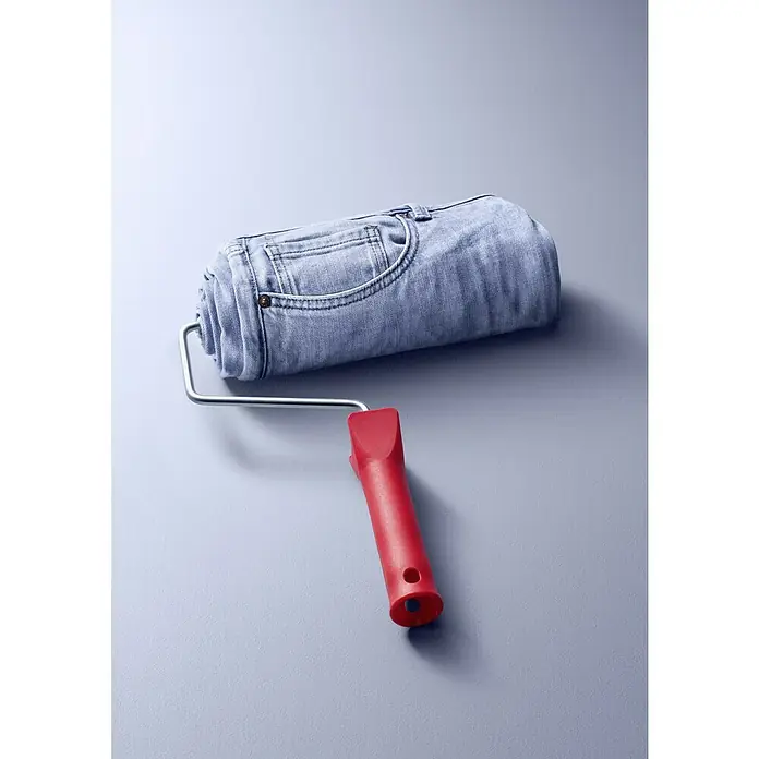 SCHÖNER WOHNEN-Farbe Wandfarbe Trendfarbe - Denim, Matt, 1 l, Konservierungsmittelfrei - Ein ungewöhnlicher Farbring: Eine zusammengerollte Jeanshose dient als "Farbwalze" auf einem Farbroller mit rotem Griff.