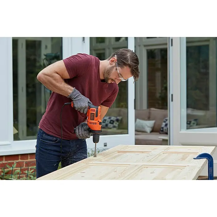 Black+Decker Taladro percutor BEH200 - 500 W, Portabrocas de sujeción rápida - Un hombre perfora una tabla de madera con un taladro Black+Decker, usando guantes y gafas de seguridad.