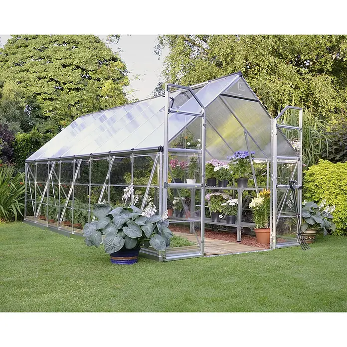 Palram – Canopia Gewächshaus Balance - Außenmaß inkl. Dachüberstand (B x T): 244 x 487 cm, 0,7 mm, Silber - Ein geräumiges Gewächshaus im Garten mit Pflanzen und Blumen in Töpfen, die auf Regalen stehen. Das Gewächshaus bietet den Pflanzen Schutz.