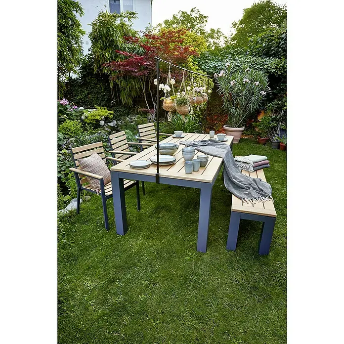 Sunfun Carina Silla de jardín - Acacia, Apilable, Natural, An x Pr x Al: 57,5 x 61 x 86 cm - Juego de mesa de jardín con tablero de madera, patas de metal azul, sillas y banco, con vajilla y decoración, en medio de un jardín exuberante.