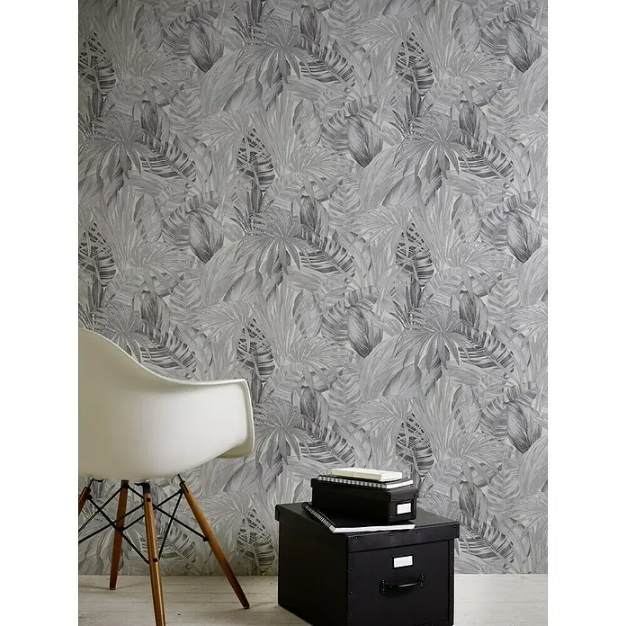 A.S. Création Greenery Vliestapete Dschungel - Schwarz/Weiß, Floral, 10,05 x 0,53 m - Nahaufnahme von einem Stuhl und einer schwarzen Box vor einer grauen tropischen Blättertapete, die eine gemütliche und stilvolle Atmosphäre schafft.