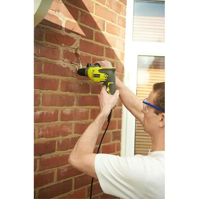Ryobi Taladro percutor RPD500-G - 500 W, Número de revoluciones de marcha en vacío 1ª marcha: 3.000 r.p.m., Número máx. de impactos: 50.000 /min - Hombre taladrando pared de ladrillo con taladro Ryobi amarillo, con gafas de seguridad.
