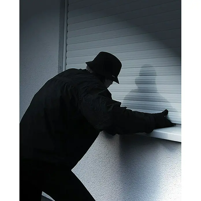 Schellenberg KlemmsicherungGeeignet für Profil-Deckhöhe: Ab 45 mm, 2 Stk. - Eine Person in dunkler Kleidung und Hut manipuliert ein Rolltor, wobei ein Schatten an der Wand sichtbar ist. Das Bild ist in gedämpften Farben gehalten.