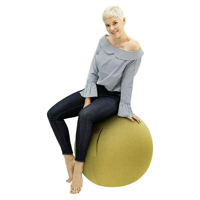 Sitting Ball Gymnastikball Felt - Senf, Durchmesser: 65 cm, 100 % Polyester - Frau sitzt auf einem gelben Sitzball, gekleidet in Jeans und einem gestreiften Hemd. Der Ball ist ein stylisches Möbelstück.