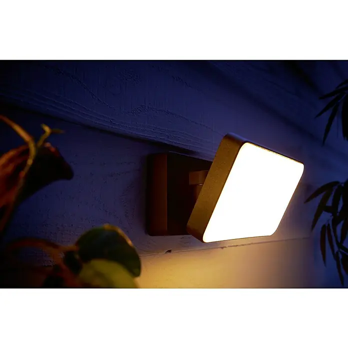 Philips Hue LED-Strahler White & Color Ambiance - 15 W, L x B x H: 22 x 16 x 15,3 cm, Schwarz, RGBW - Nahaufnahme einer modernen, quadratischen Außenwandleuchte, die ein helles, warmes Licht an eine blaue Wand wirft.