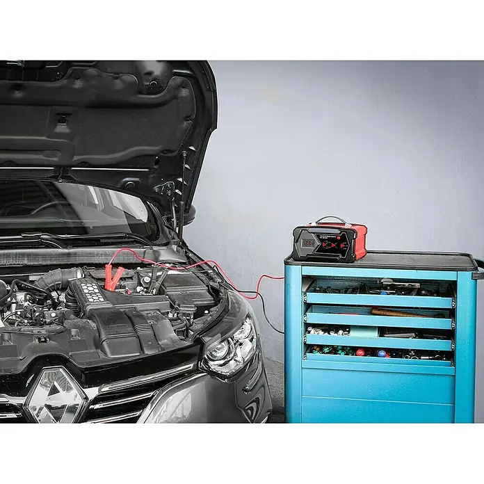 APA Cargador para el taller - Corriente de carga: 2 A - 12 A - Cargador de batería de coche junto a un coche con el capó abierto. El cargador rojo está sobre un carro de herramientas azul.