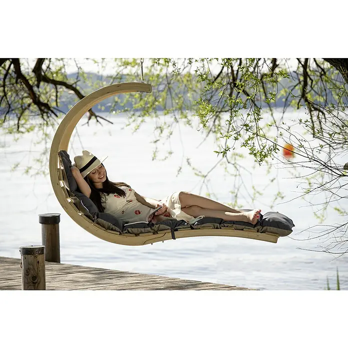 Amazonas Hängeliege Swing Lounger - L x B x H: 240 x 75 x 114 cm, Anthrazit - Eine Frau entspannt sich auf einer Hängeliege aus Holz mit grauen Kissen auf einem Steg mit Blick auf das Wasser.