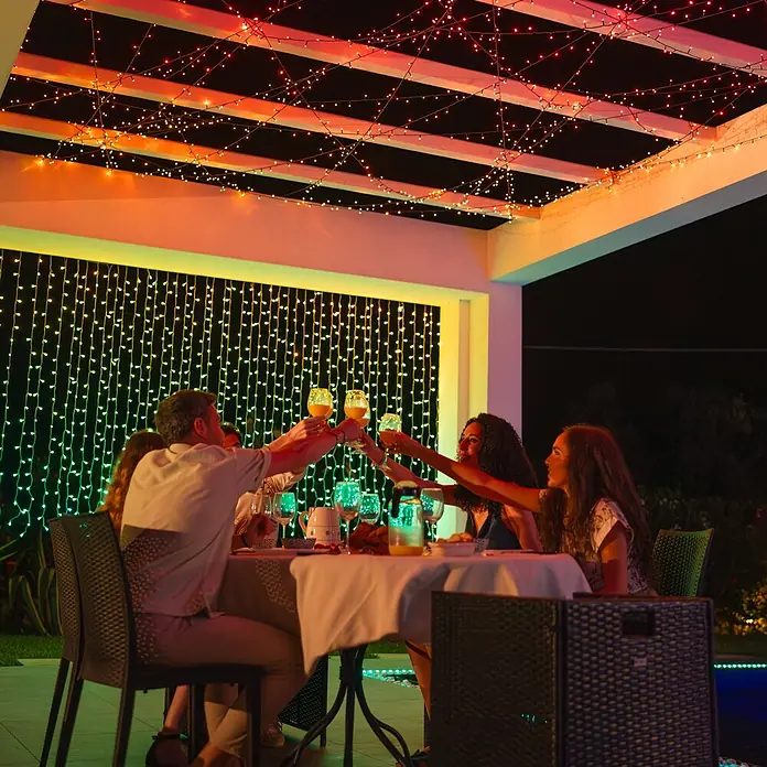 Twinkly Smart-LED-Lichterkette StringsAußen, 20 m, Lichtfarbe: RGBW, Anzahl Leuchtmittel: 250 Stk., Netzbetrieben - Abendliche Feier mit farbigen Lichtern. Eine Gruppe von Menschen stößt mit Getränken an einem Tisch unter funkelnden Lichtern an.