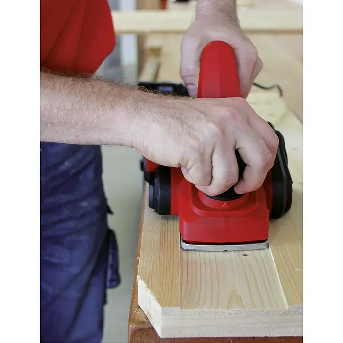 Einhell Elektrohobel TC-PL 750 - 750 W, Hobeltiefe: 0 mm - 2 mm, Leerlaufdrehzahl: 0 U/min - 17.000 U/min - Bild von einer Person, die mit einem roten Elektrohobel ein Stück Holz bearbeitet. Der Hobel ist das Hauptaugenmerk des Bildes.
