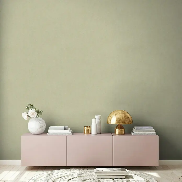 A.S. Création New Walls Vliestapete Textil - Hellgrün, Uni, 10,05 x 0,53 m - Dieses Bild zeigt ein stilvolles rosa Sideboard mit Dekor wie einer goldenen Lampe, Büchern und einer Vase mit Blumen vor einer grünen Wand.