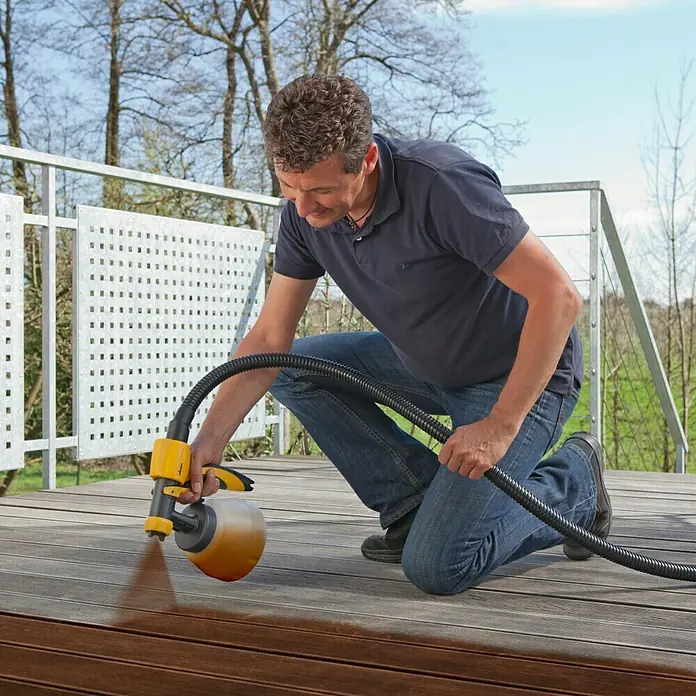 Wagner Sprühaufsatz Wood & Metal EXTRA Large - Geeignet für: Dünnflüssige lösemittel- & wasserbasierte Materialien - Ein Mann kniet auf einem Holzdeck und sprüht es mit einer gelben Farbspritzpistole. Perfekt zum einfachen Auftragen von Farbe im Außenbereich.