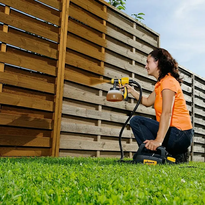 Wagner Sprühaufsatz Wood & Metal EXTRA Large - Geeignet für: Dünnflüssige lösemittel- & wasserbasierte Materialien - Eine Frau sprüht mit einer Wagner Farbspritzpistole einen Holzzaun, um ihn zu schützen und zu verschönern.