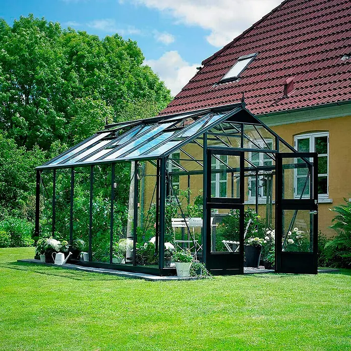 Juliana Gewächshaus Premium - Außenmaß inkl. Dachüberstand (B x T): 296 x 439 cm, Material Seitenwand: Glas, 3 mm, Anthrazit/Schwarz - Ein elegantes Gewächshaus aus Glas und dunklem Rahmen steht im Garten. Die Tür ist offen und zeigt Pflanzen im Inneren.