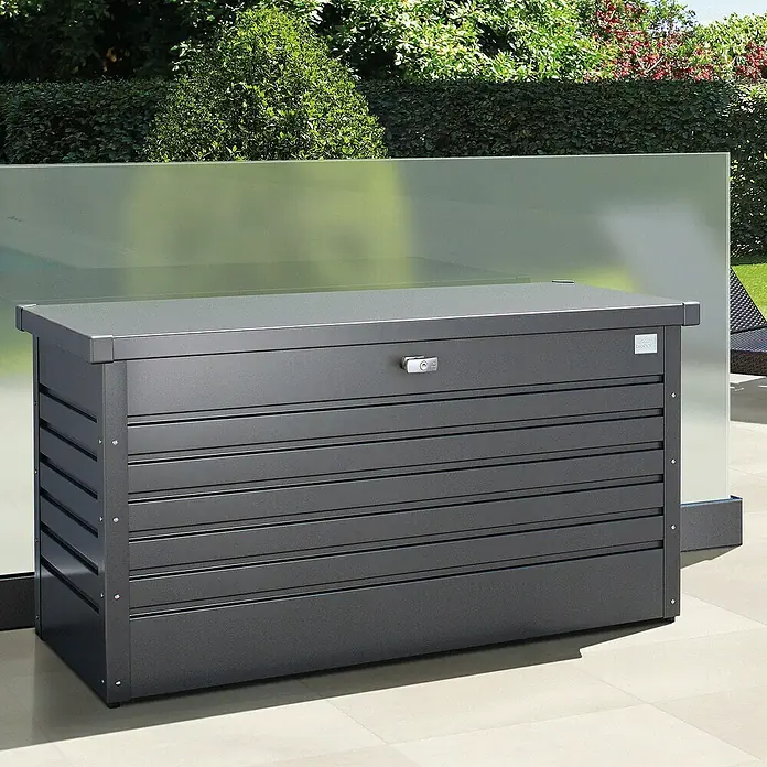 Biohort FreizeitBox Gartenbox FreizeitBox 130 - L x B x H: 134 x 62 x 71 cm, Dunkelgrau Metallic, Stahl - Eine graue Biohort-Aufbewahrungsbox aus Metall steht auf einer Terrasse vor einer Glaswand und Grünpflanzen.