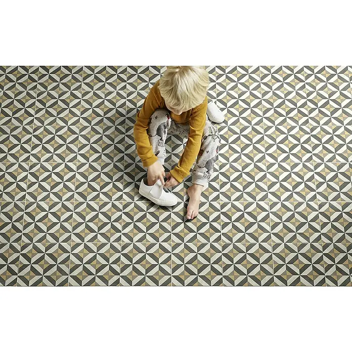 Beauflor PVC-Boden Meterware Orion400 cm, Tile 04, Olivgrün/Beige - Bild eines Kindes, das auf einem Boden mit geometrischen Fliesen sitzt und weiße Schuhe anzieht.