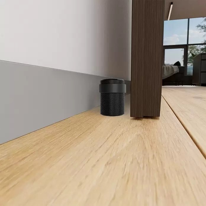 Walteco Bodentürstopper - Ø x H: 34 x 40 mm, Metall - Schwarzer, zylindrischer Türstopper mit Textur und Gummiband, auf Holzboden zwischen Tür und heller Wand.