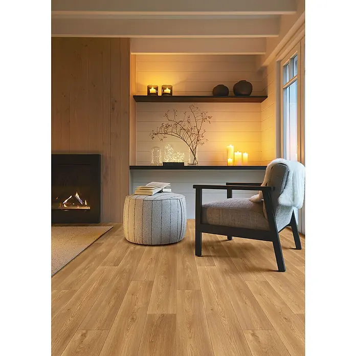 Beauflor PVC-Boden Meterware Themis - Wood 02, Breite: 400 cm, Braun - Gemütliche Leseecke mit einem bequemen Sessel und einem runden Hocker auf einem hellen Holzboden. Der Raum wird durch ein Kaminfeuer und Kerzen beleuchtet.