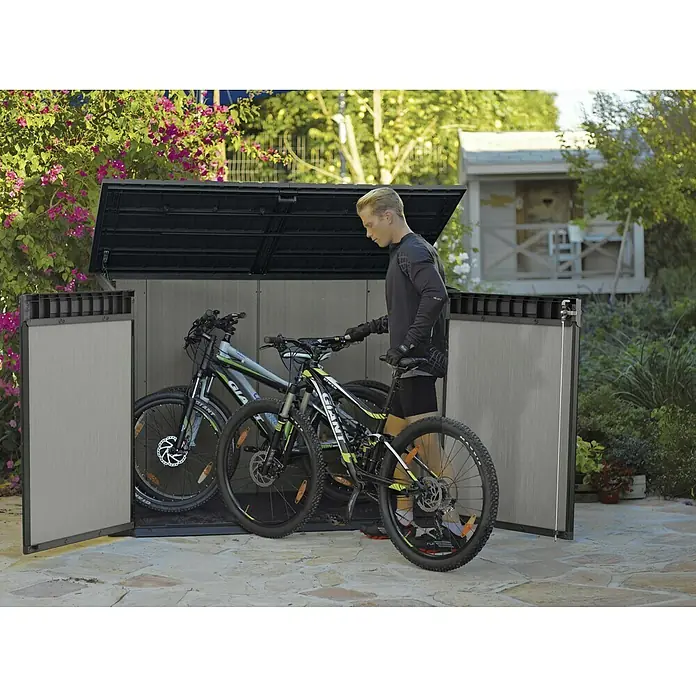 Keter Truhe Grande Store - Außenmaß inkl. Dachüberstand (B x T): 191 x 109 cm, Kunststoff, Grau, Geeignet für Anzahl Mülltonnen: 3 Stk. - Ein Mann schiebt ein Fahrrad in eine abschließbare Fahrradbox aus Kunststoff im Freien. Stauraumlösung für Fahrräder.