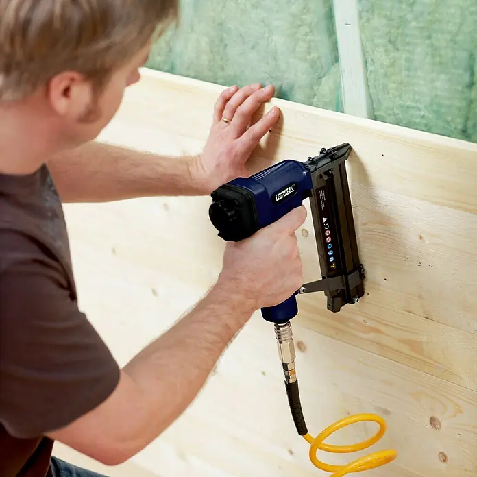 Rapid Kombidrucklufttacker AirTac PBS 151 - Länge Nagel: 15 mm - 50 mm, Geeignet für: Nageltyp 8, Pneumatisch - Nahaufnahme eines Mannes, der einen Rapid Druckluftnagler verwendet, um Holz zu befestigen. Das Werkzeug ist blau mit einem gelben Schlauch.